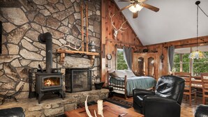 A cozy cabin interior with a stone fireplace, a bed, leather furniture, and a large window overlooking greenery. - Rustic retreat w/ kitchen & deck (Great Cacapon)