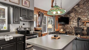 A modern kitchen with dark cabinetry, a large island, a stone accent wall, and a hanging light fixture. - Rustic retreat w/ kitchen & deck (Great Cacapon)
