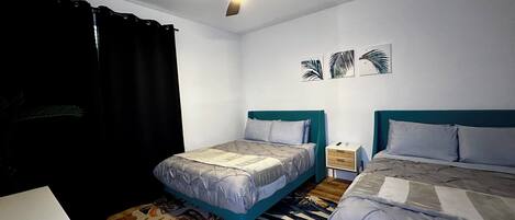 Une chambre avec deux lits, un ventilateur de plafond, une table de chevet et un mur orné de trois tableaux représentant des motifs de feuilles.