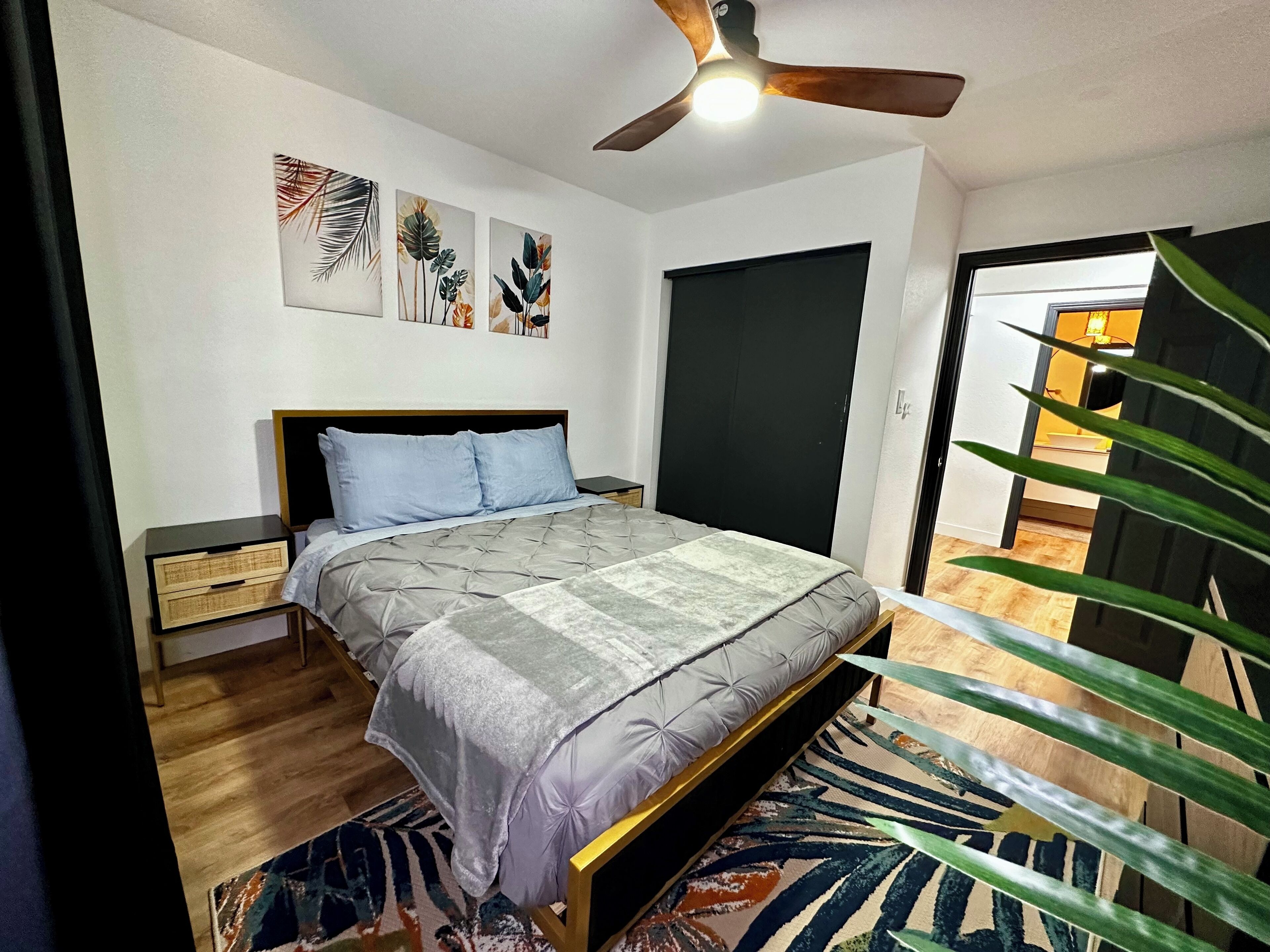 A bedroom with a bed, a ceiling fan, a nightstand, a door, and a plant.