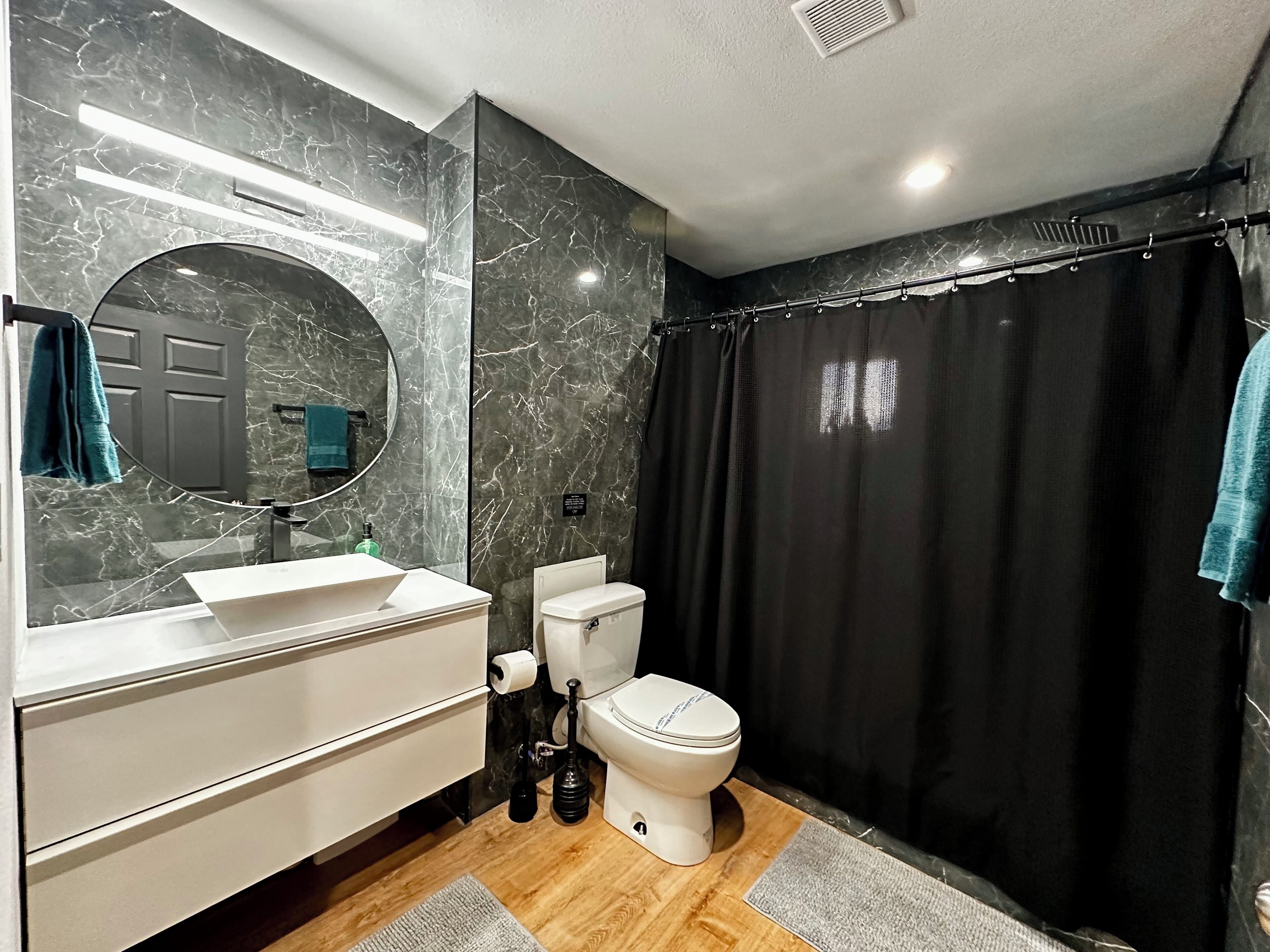 Une salle de bain moderne avec un pare-baignoire en marbre, un miroir rond, un lavabo blanc, des toilettes et un rideau de douche noir.