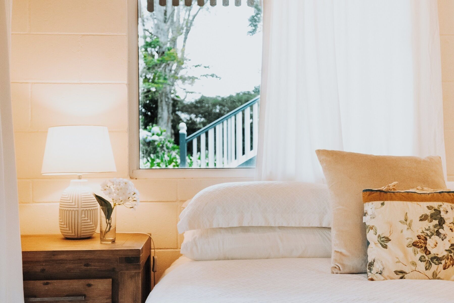 A bedroom with a bed, a nightstand with a lamp, and a window with a view of a balcony and trees.