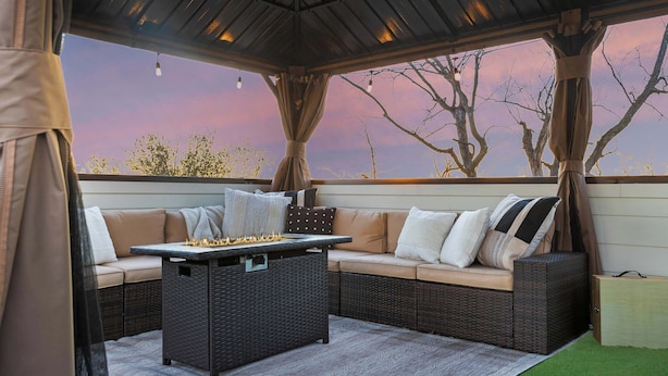 An outdoor seating area with a table and cushions, under a covered structure with a view of trees and a sunset. - Lyric | Luxury Stay with Rooftop & Firepit (Nashville)