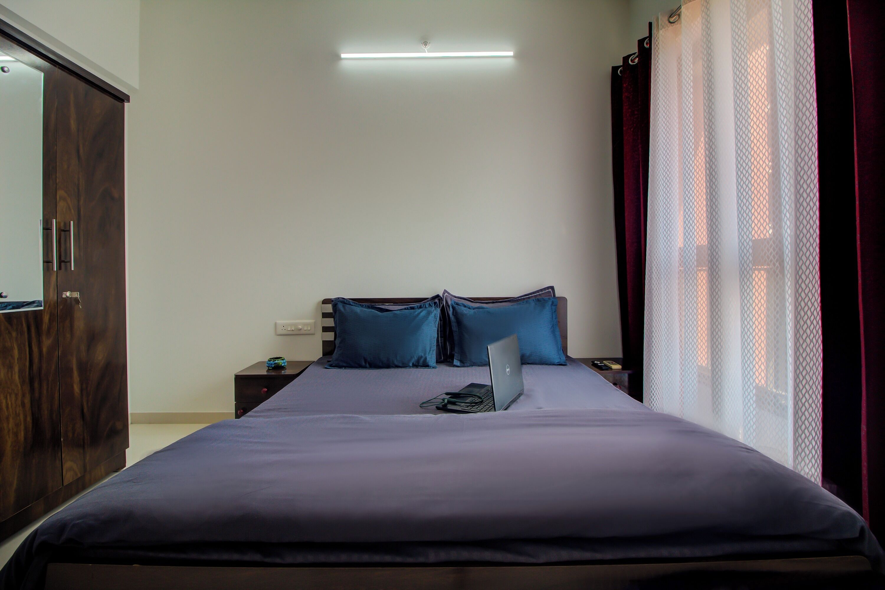 Una cama bien hecha con una laptop, un armario de madera y una mesita de noche con un teléfono.