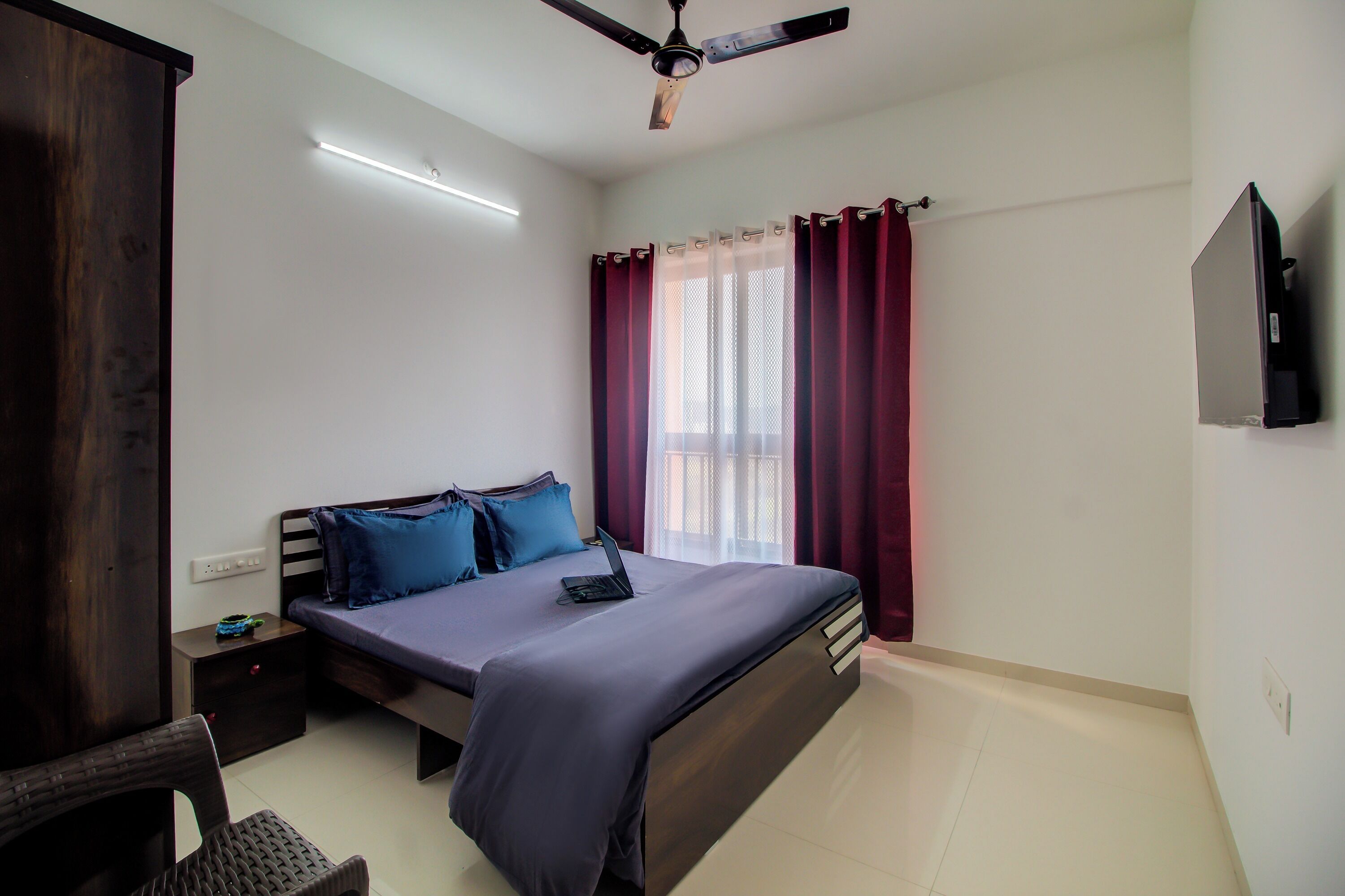 A bedroom with a bed, a desk, a chair, a ceiling fan, and a TV on the wall.