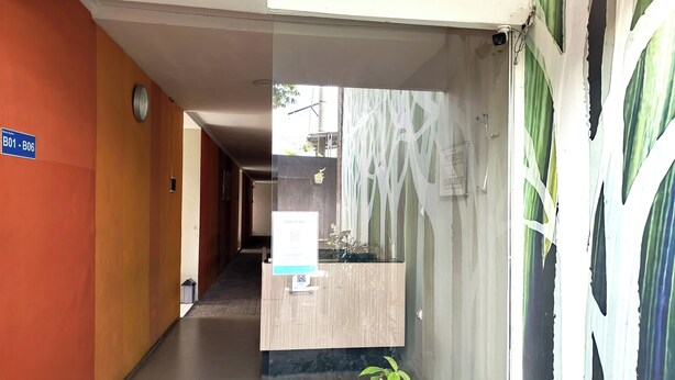 A hotel reception area with a wooden reception desk, a clock on the wall, and a sign indicating room numbers B01-B06. - Khanza Costel Syariah (Karangtengah)