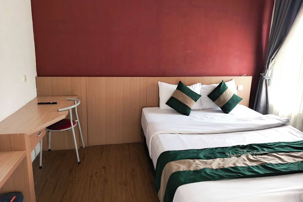 Una habitación de hotel con un escritorio de madera, una cama con almohadas verdes y beige, y una pared de acento roja.