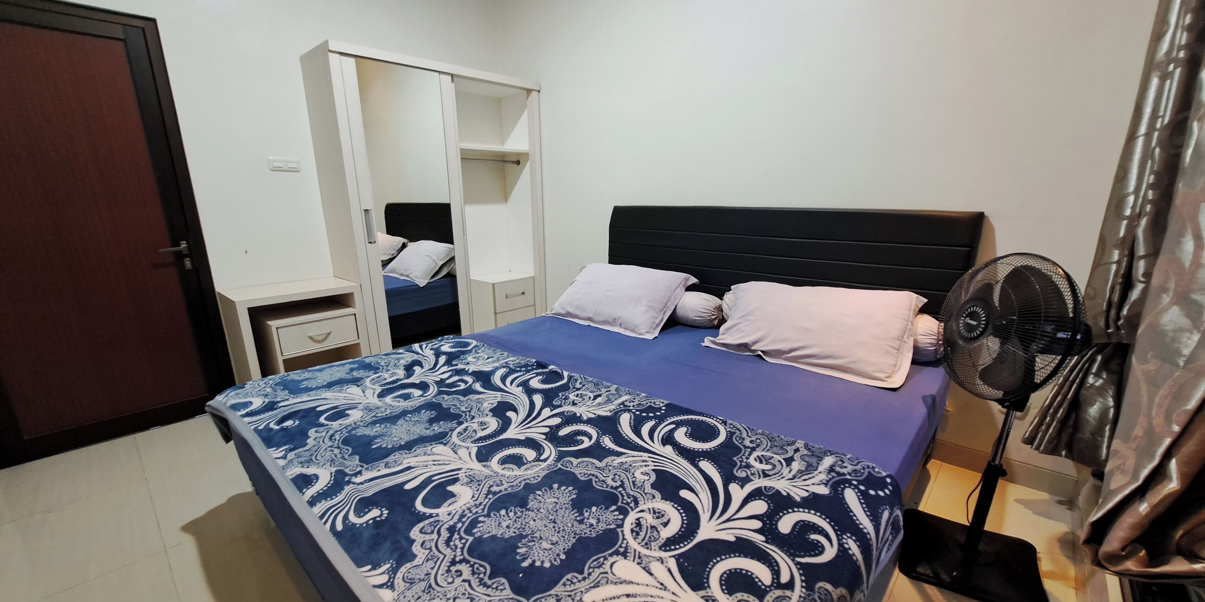 A bedroom with a bed, a wardrobe, a bedside table, and a standing fan.