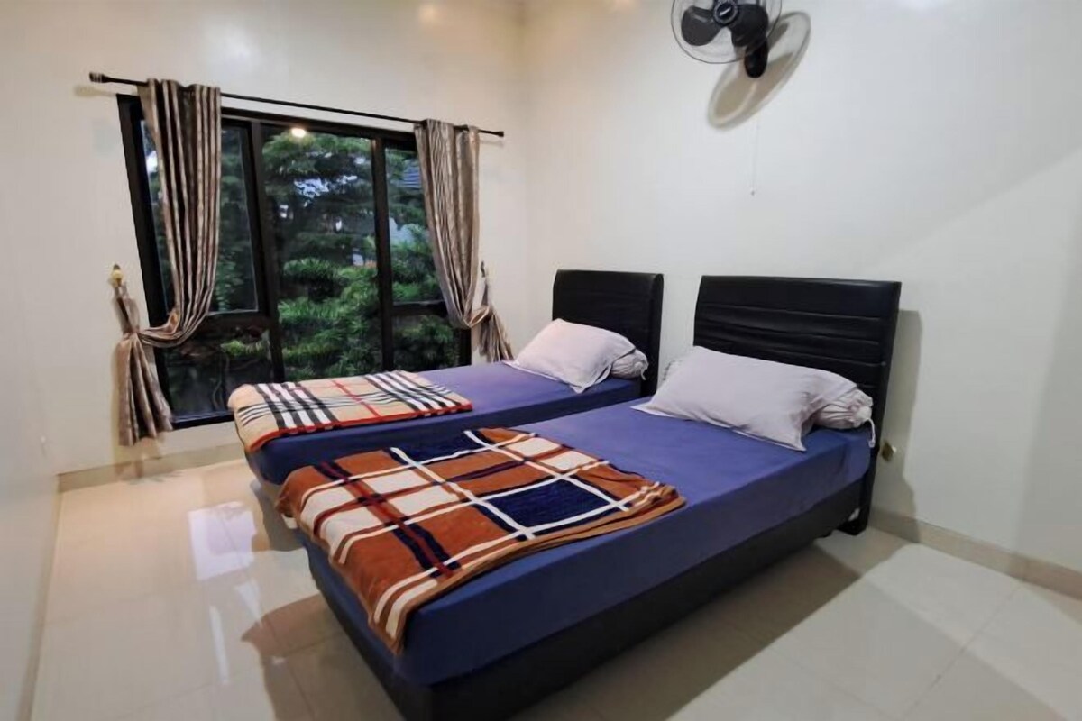 Una cama doble con una colcha azul y una manta colorida, una ventana con cortinas y un ventilador de pared.