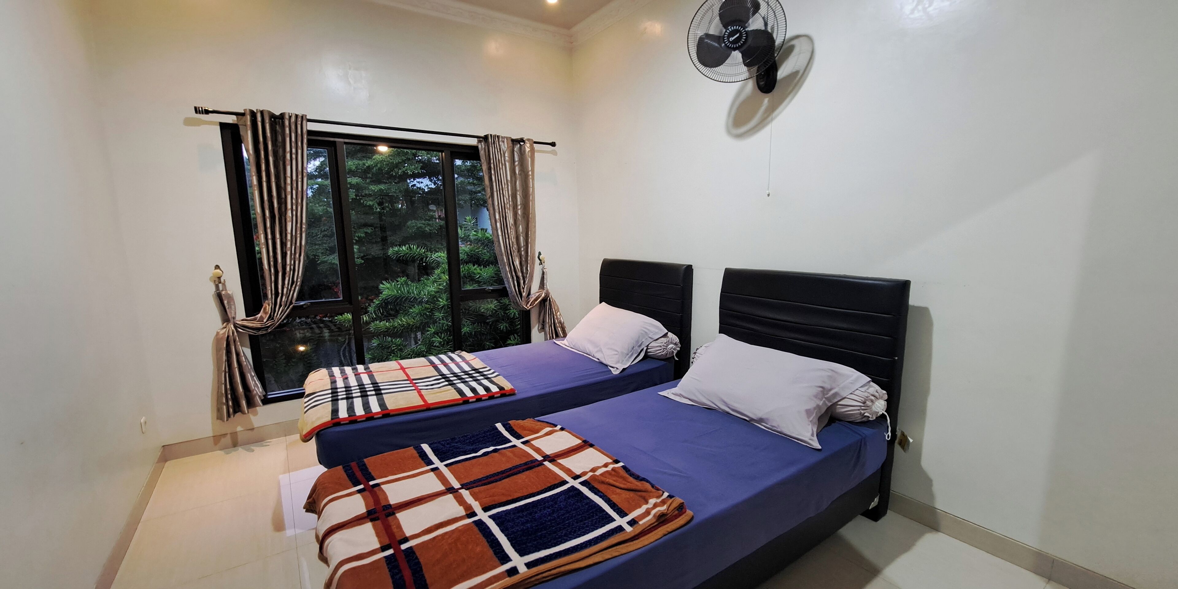 A bedroom with two beds, a window with curtains, a ceiling fan, and a view of greenery outside.