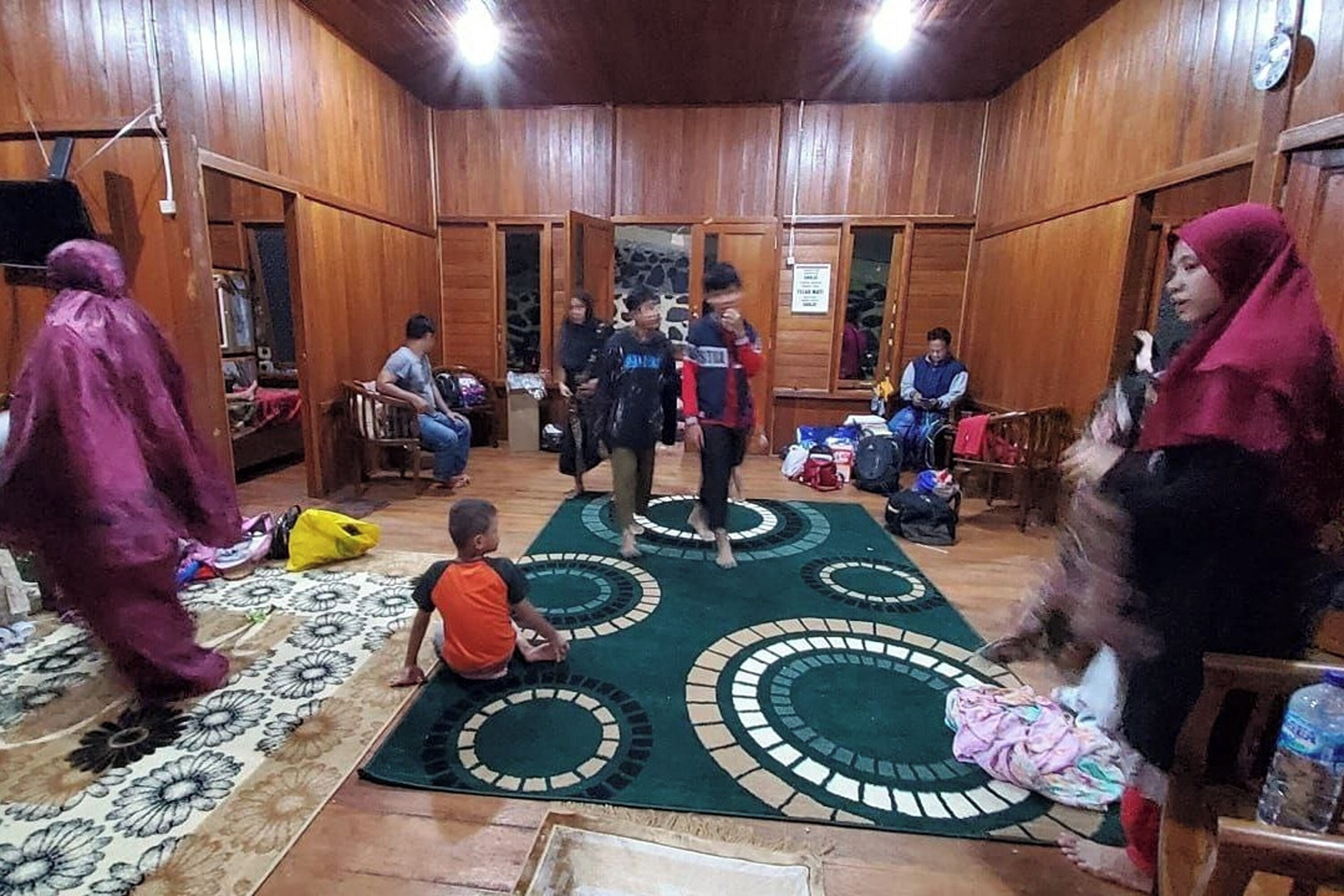 A room with wooden walls and a green carpet with circular patterns. There are several people, some standing and some sitting, and a child playing on the floor.