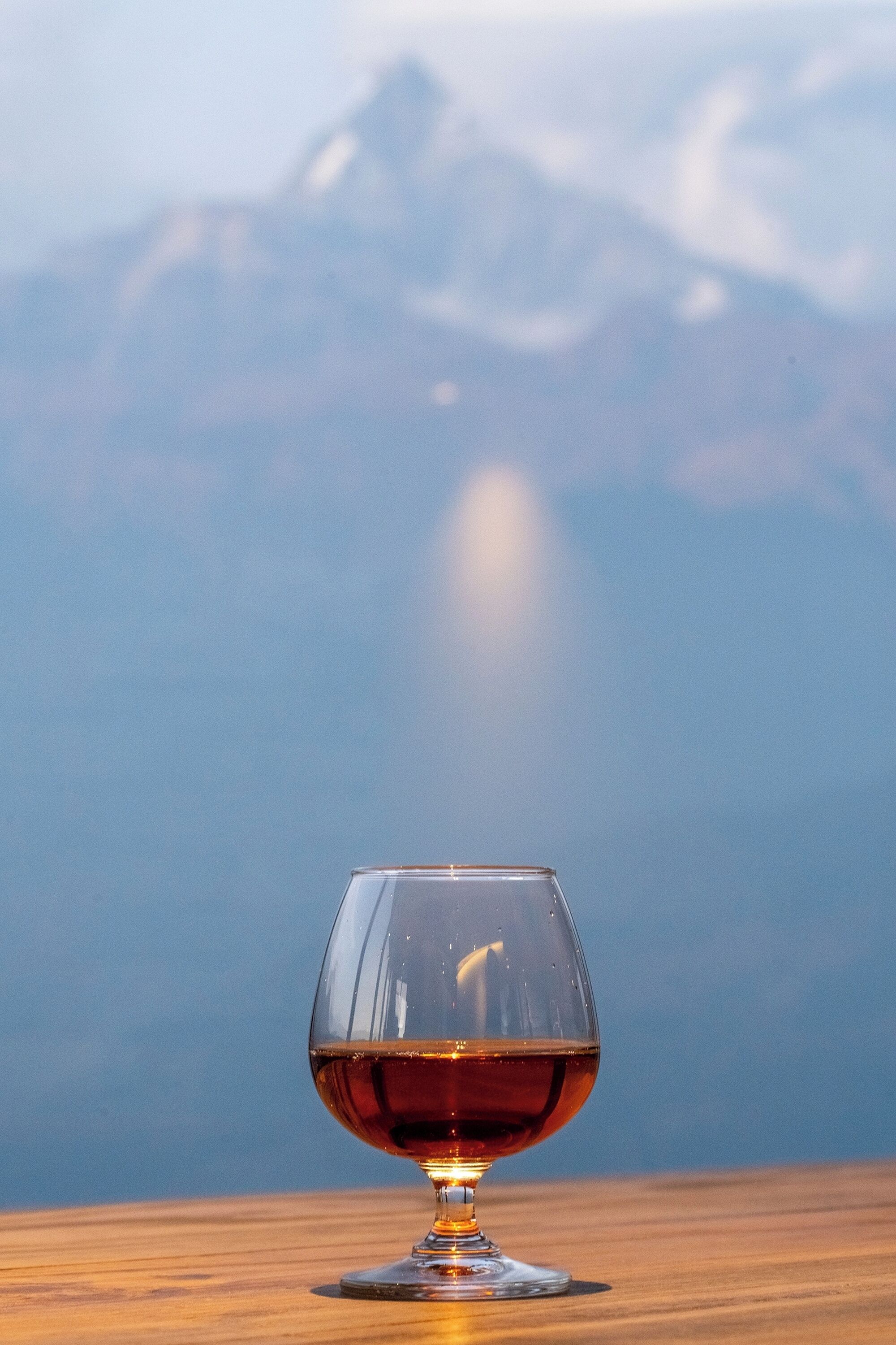 Ein klares Glas mit bernsteinfarbener Flüssigkeit auf einer Holzoberfläche, im Hintergrund eine Berglandschaft.