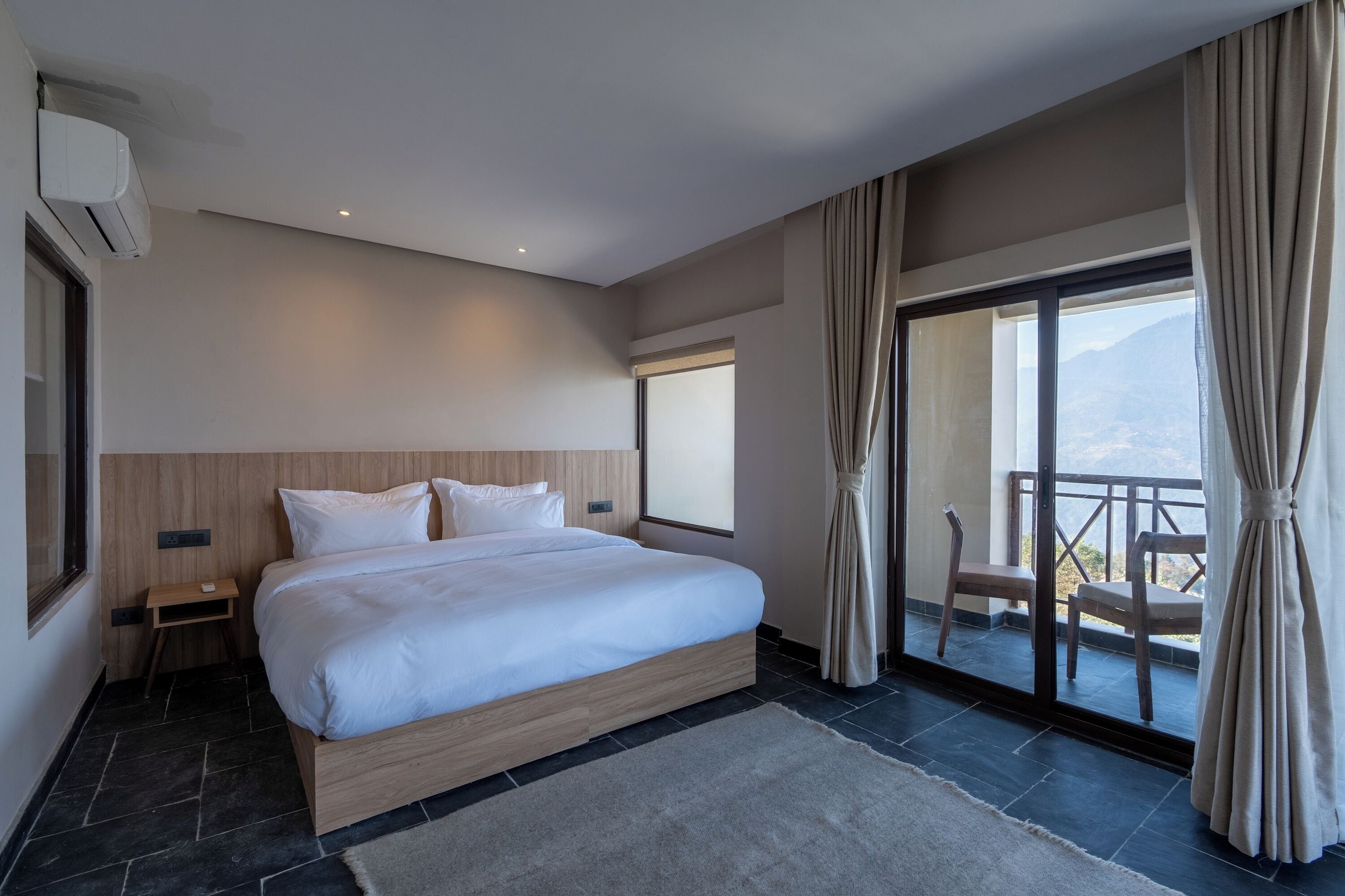 A modern hotel room with a large bed, a small bedside table, a window with curtains, and a balcony with chairs.