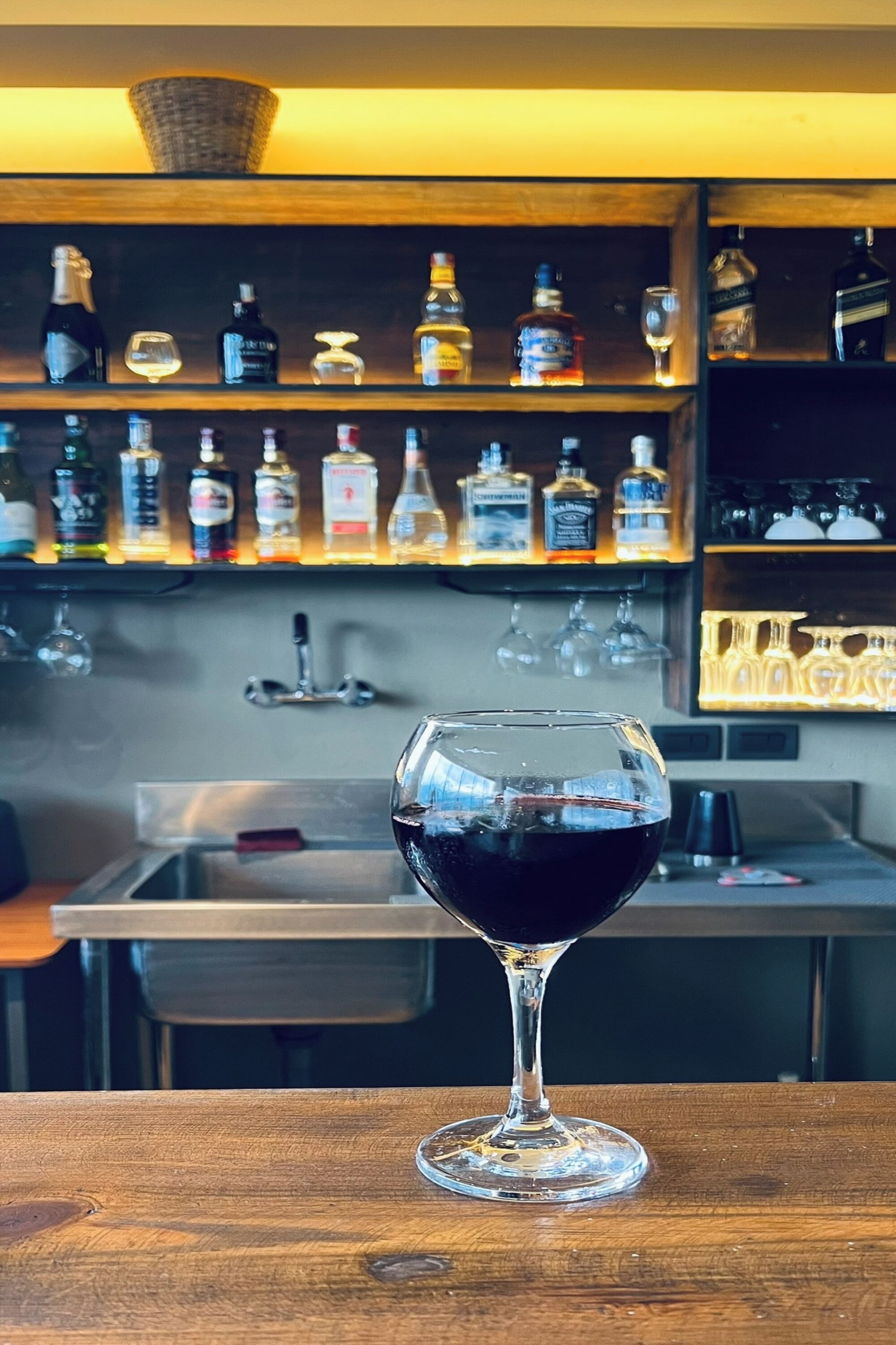 A bar area with a glass of red wine on a wooden table, shelves with various bottles and glasses, and a bar sink.