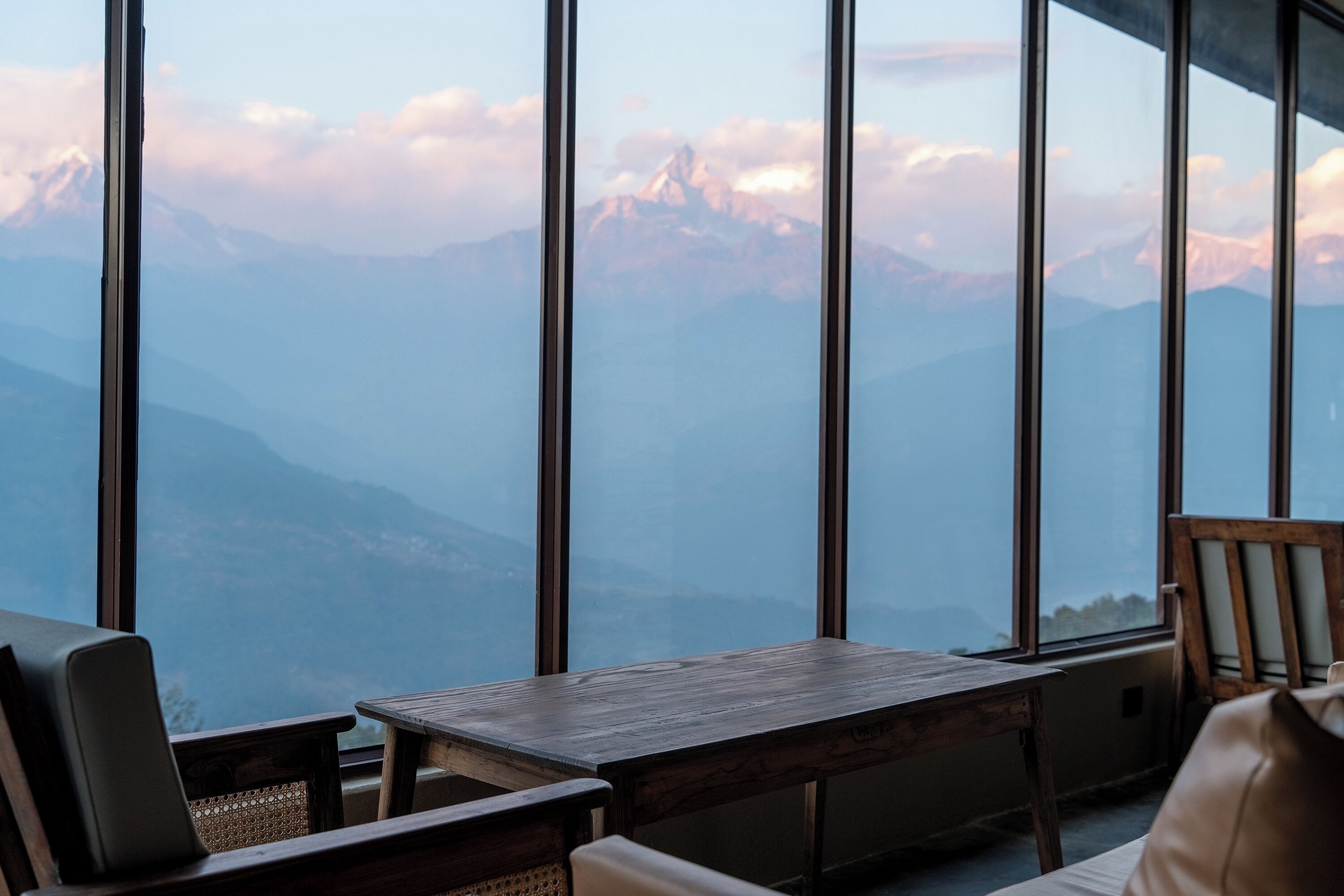 A room with a large window offering a mountain view. There is a wooden table and two chairs.