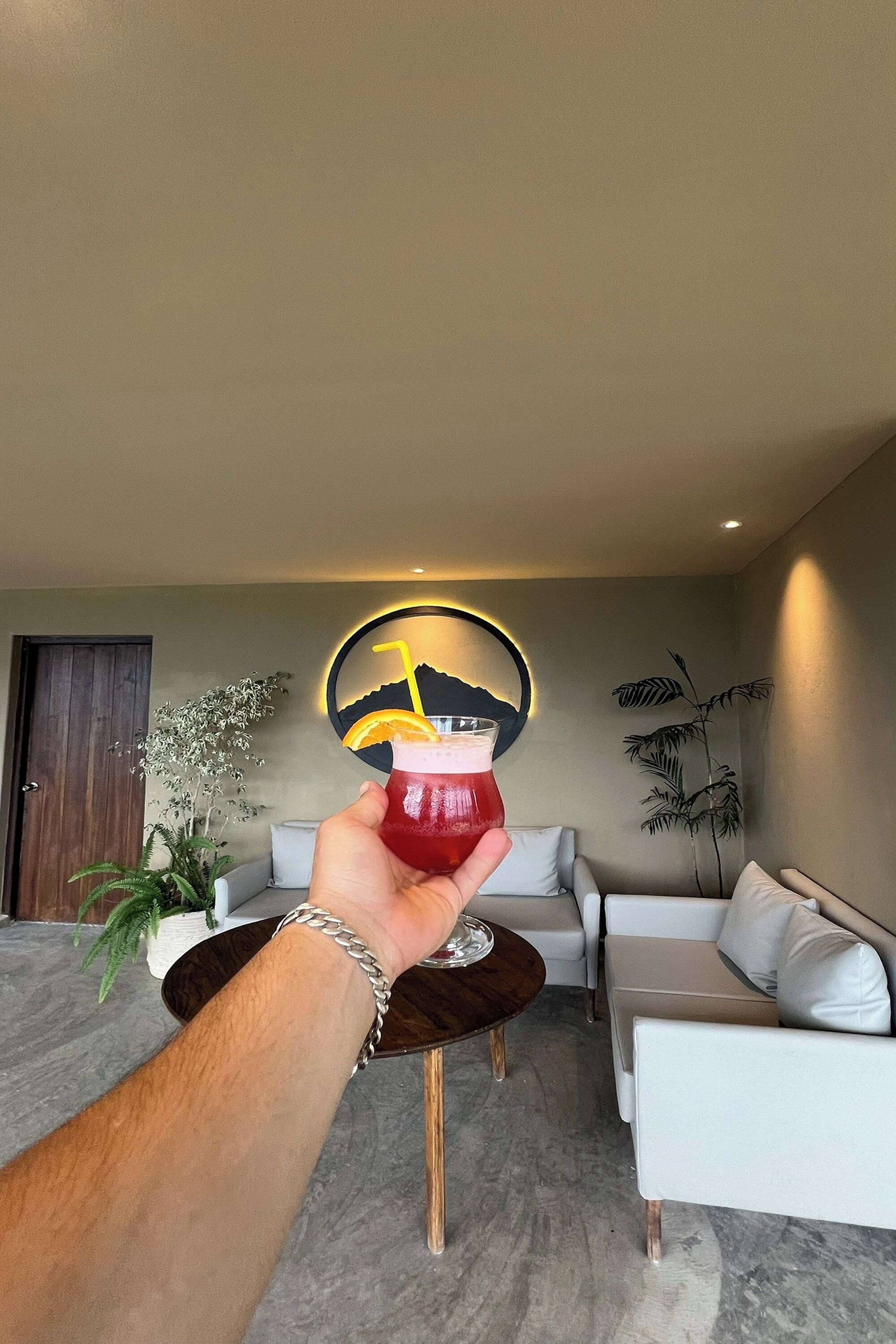 A hand holding a drink in a modern living room with a sofa, a coffee table, and a wall art piece.