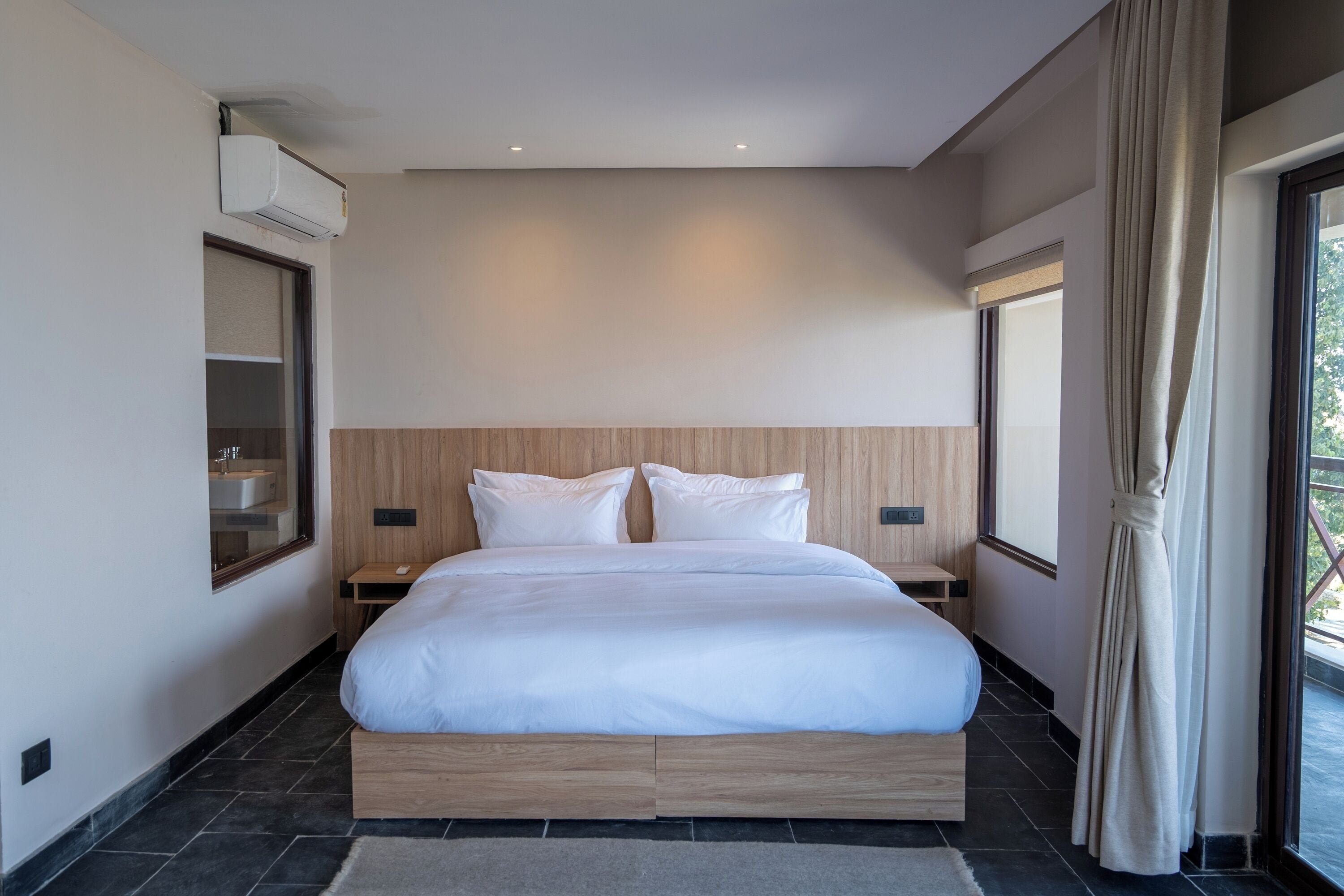A modern bedroom with a large bed, built-in nightstands, a wall-mounted air conditioner, and a window with curtains.