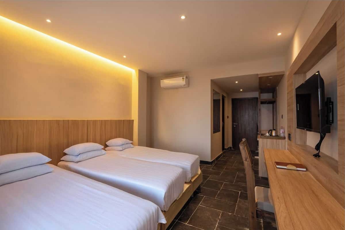 A hotel room with three beds, a wooden headboard, a flat-screen TV mounted on the wall, and a small kitchenette area.