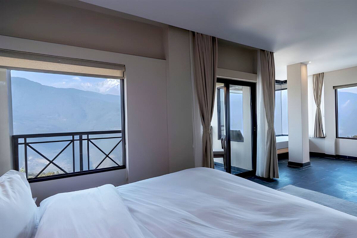 A hotel room with a large bed, a balcony with a view of mountains, and a sliding door leading to another room.