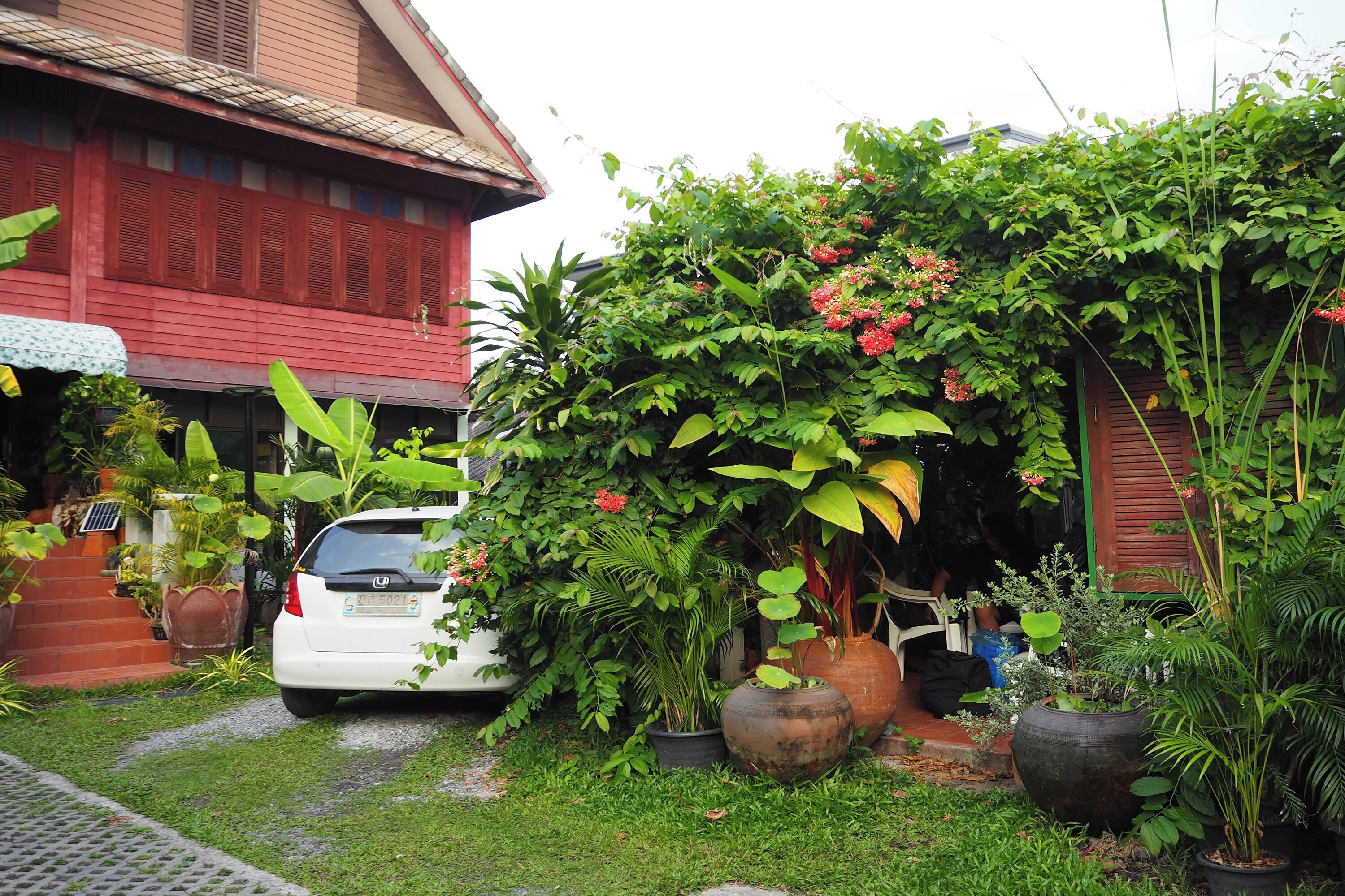 A white car parked in front of a red building with a garden area containing various potted plants and a small staircase leading to the entrance.