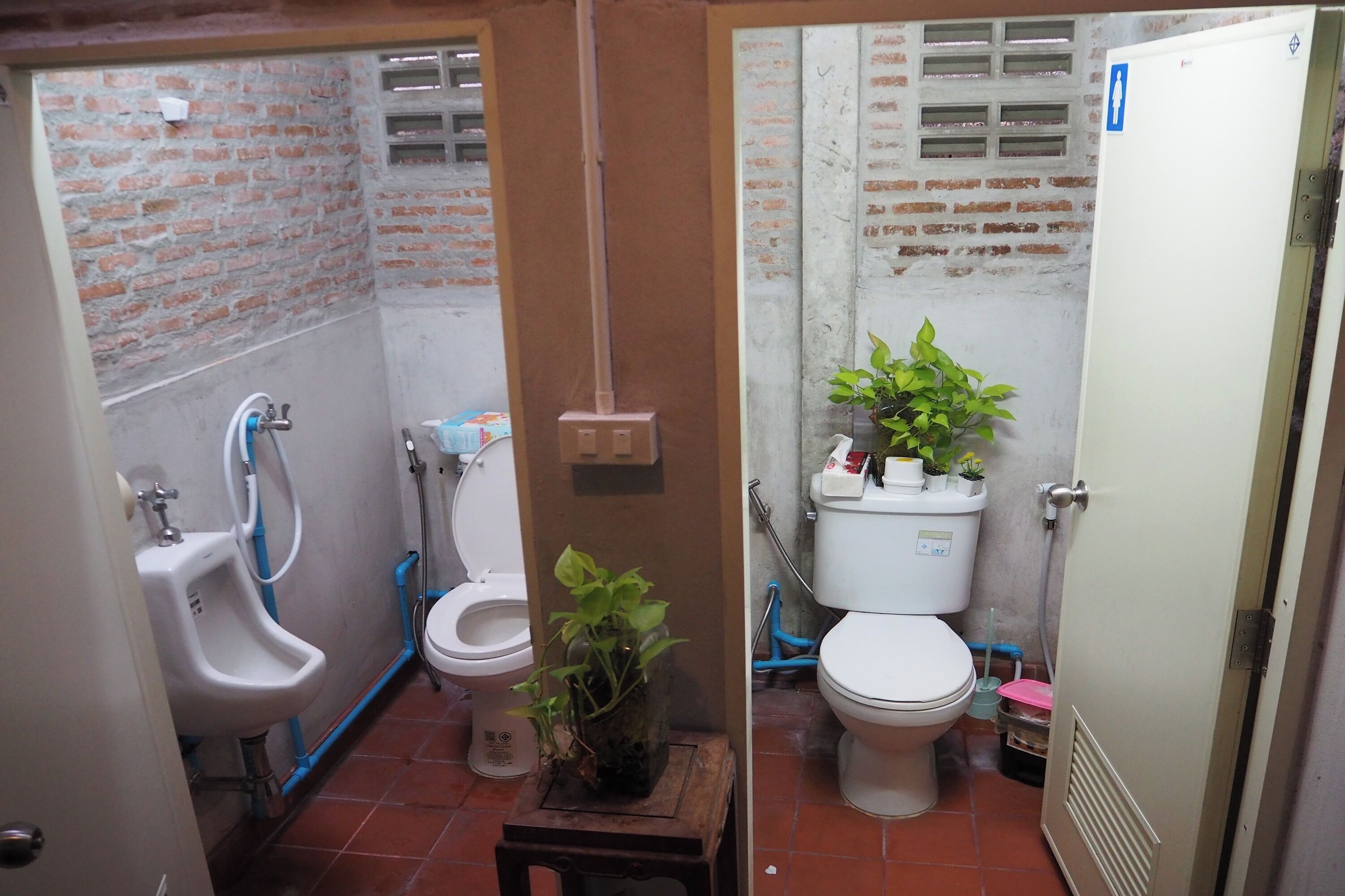 Une petite salle de bain compacte, équipée d’un urinoir, de toilettes et d’un lavabo. Une plante en pot est posée sur une petite table, et les murs sont en briques.