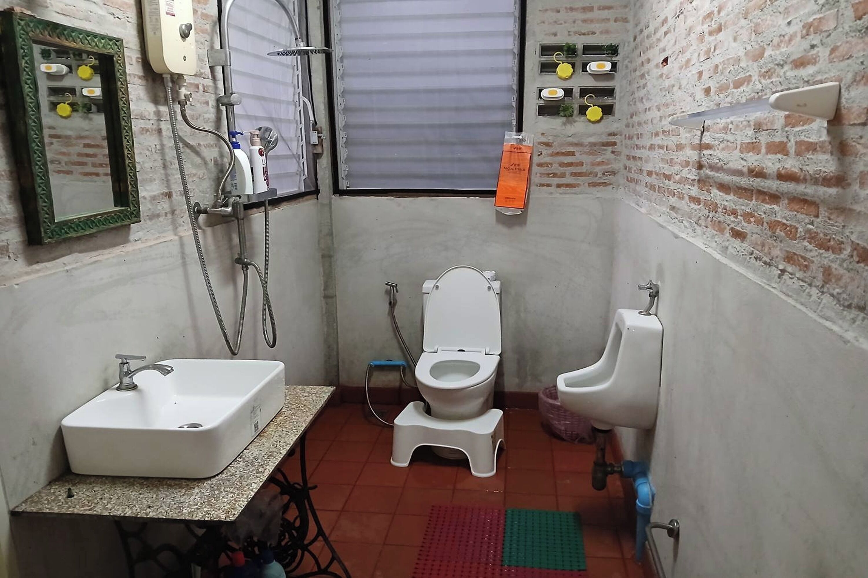 A compact bathroom with a sink, toilet, and urinal, a mirror, and a shower with a hose.