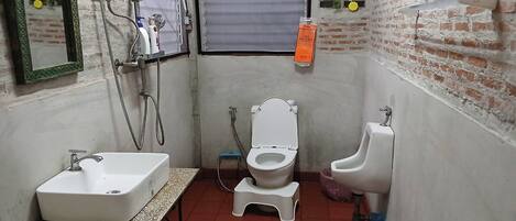 A compact bathroom with a sink, toilet, and urinal, a mirror, and a shower with a hose.
