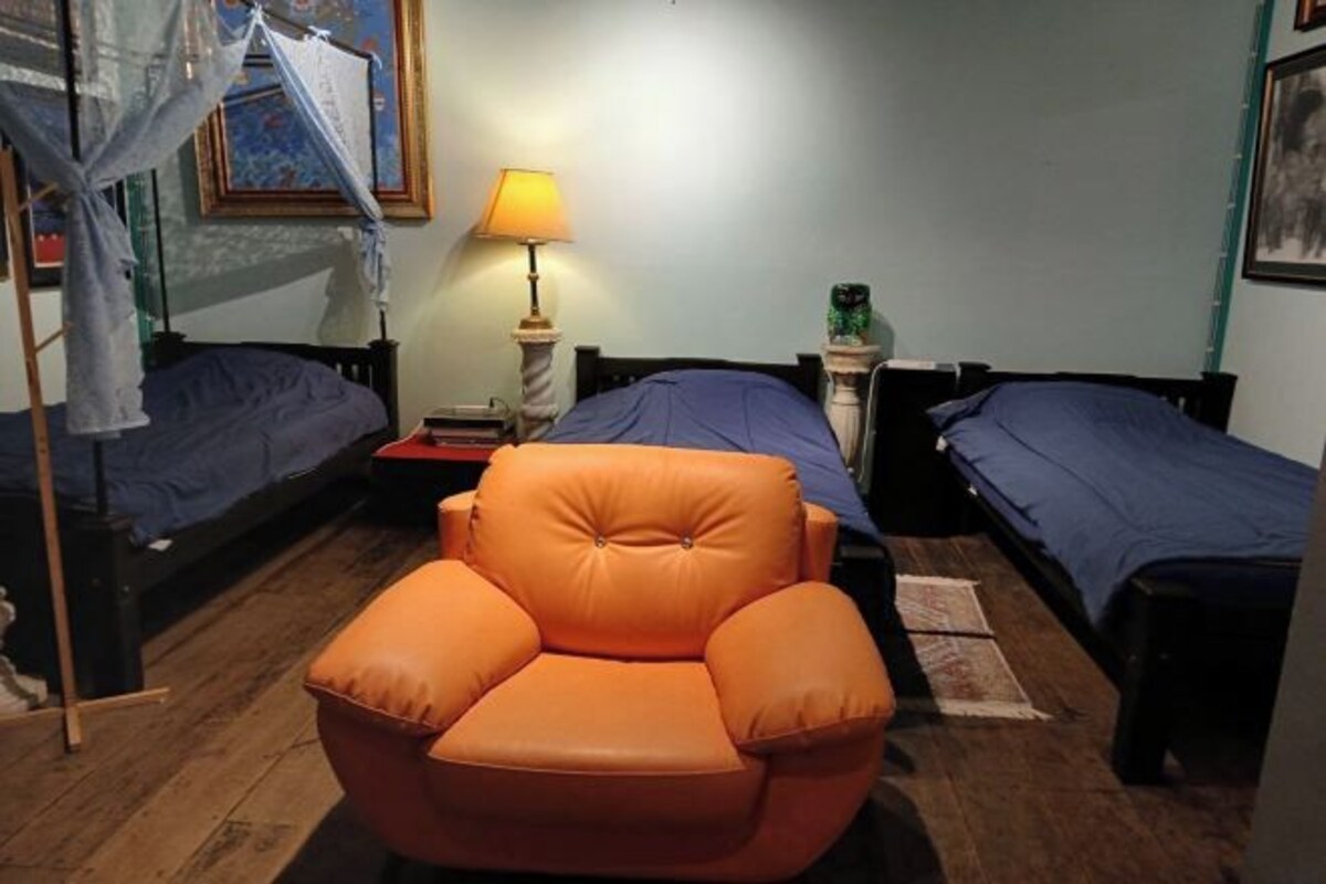 A room with two single beds, a wooden floor, a chair, a nightstand with a lamp, and a picture frame on the wall.