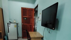 Een kleine, goed verlichte kamer met een houten bureau, een aan de muur bevestigde televisie, een koelkast, en een deur met een nummerbord.