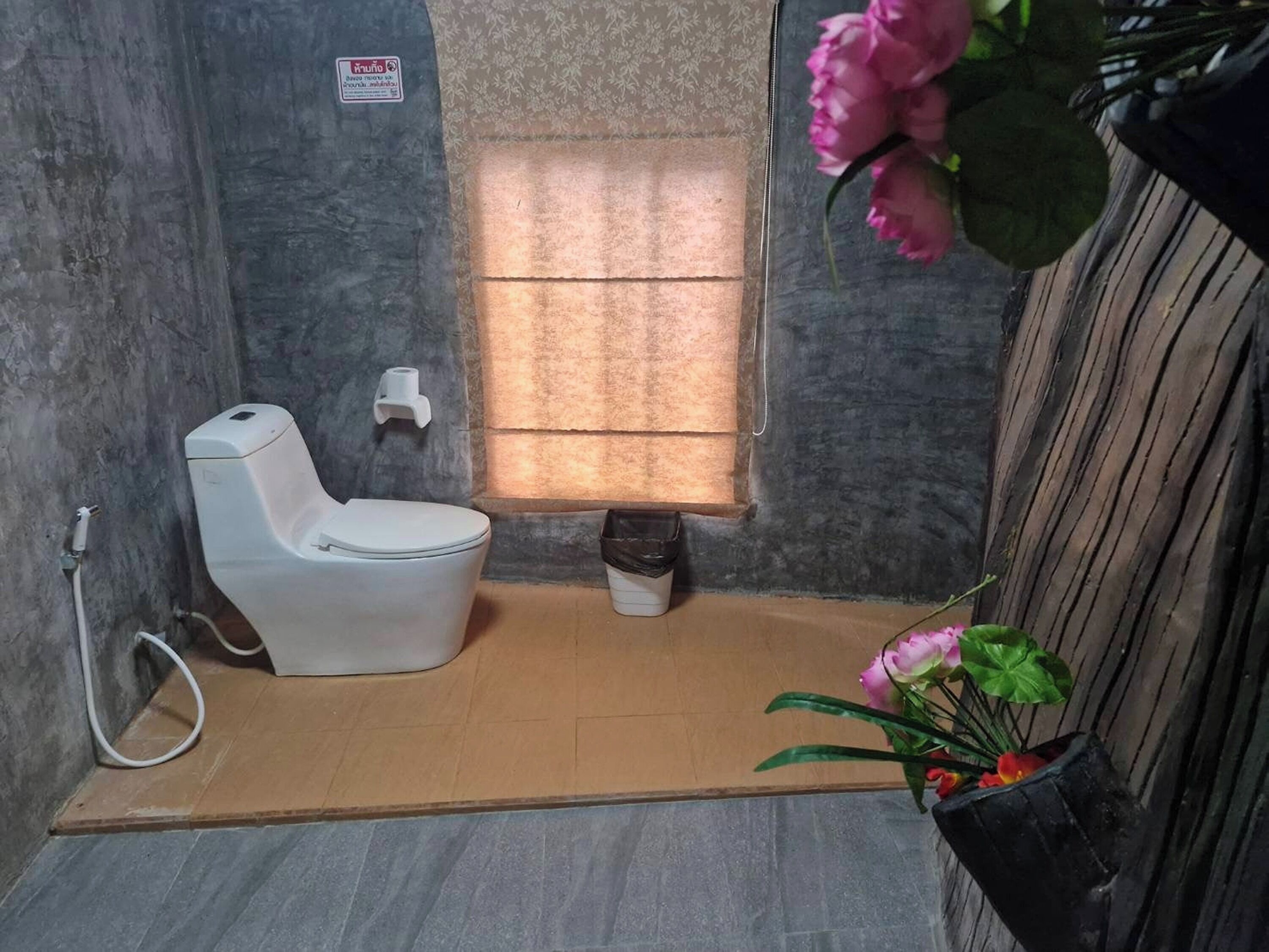 Un baño pequeño con inodoro, ventana con cortinas, un cubo de basura y una planta en maceta.