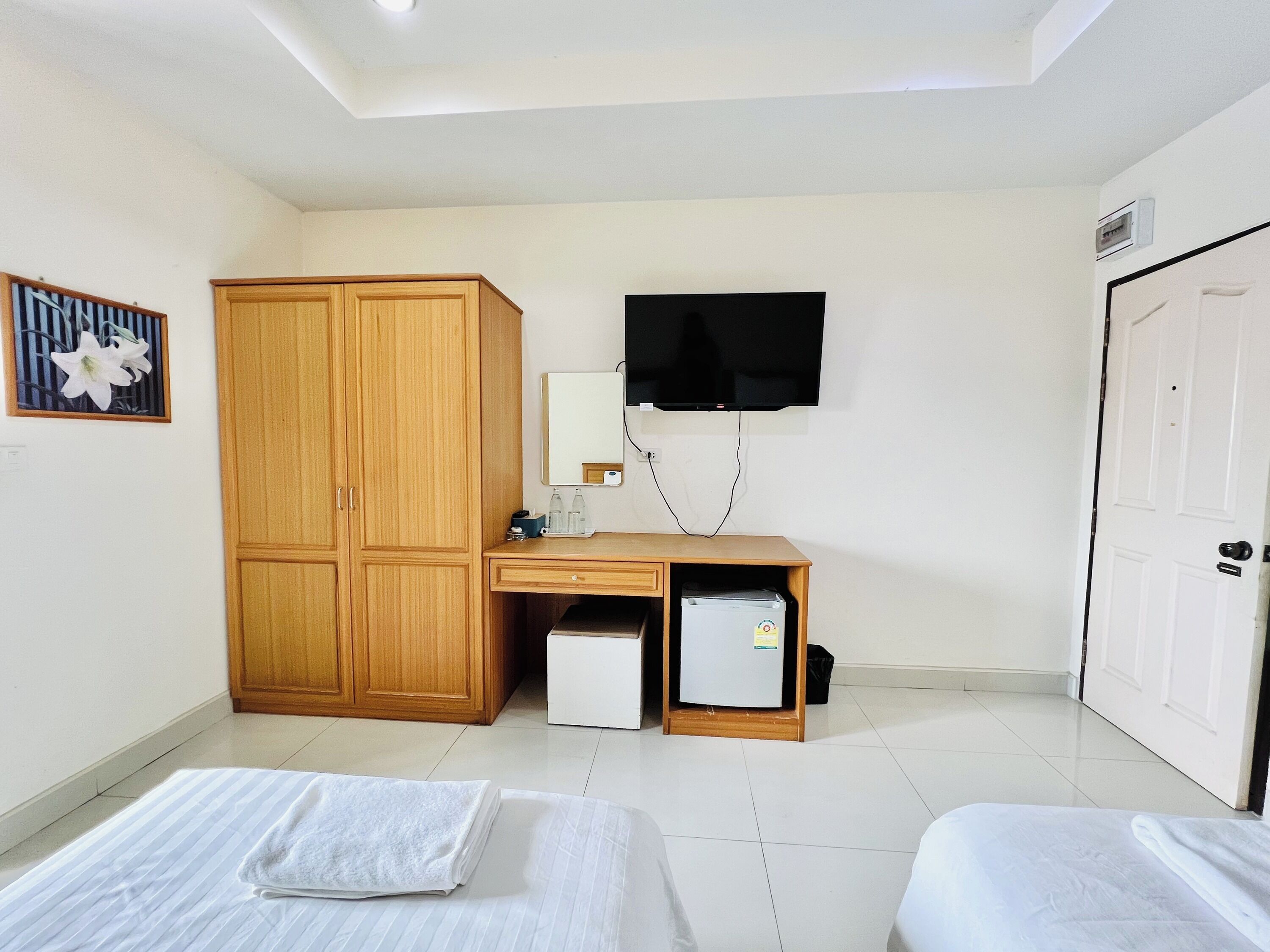A hotel room with a wooden wardrobe, a small fridge, a desk with a mirror, and a TV mounted on the wall.