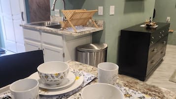 A kitchen with a sink, a wooden drying rack, a black cabinet, and a dining table set with floral-patterned plates and cups.