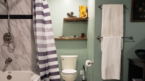 A bathroom with a bathtub, toilet, and shower curtain. There are two rugs on the floor and a shelf with decorative items above the toilet. - Mountain Chic Cabin Retreat (Woodland Park)