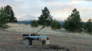 A concrete structure with a shovel in a grassy field with trees and mountains in the background. - Mountain Chic Cabin Retreat (Woodland Park)