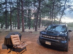 A black SUV parked in a forested area with a camping chair and a tent nearby. - Mountain Chic Cabin Retreat (Woodland Park)