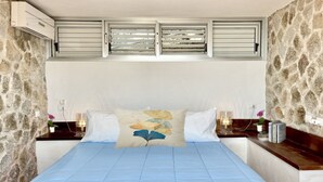 Ein Schlafzimmer mit Steinstruktur, einem Bett mit blauer Bettdecke, einem Zierkissen, Nachttischlampen und einem kleinen Tisch mit Büchern und einer Pflanze.
