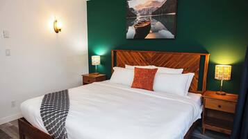 A neatly made bed with white linens and a patterned throw, flanked by wooden nightstands with lamps, in a room with green walls and a framed picture above the bed.