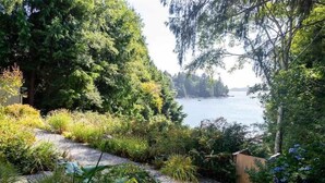 A garden with a gravel path leading to a body of water, surrounded by dense foliage and a wooden structure. - Tofino Oceanfront Hotel Suite (Tofino)