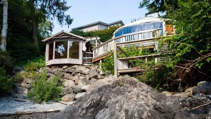 A wooden cabin with a deck, surrounded by rocks and vegetation, with a house in the background. - Tofino Oceanfront Hotel Suite (Tofino)