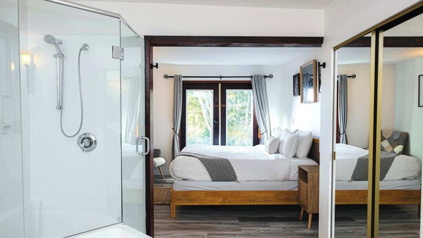 A modern bedroom with a glass-enclosed shower, a wooden bed with white bedding, and a large mirror reflecting the room. - Tofino Oceanfront Hotel Suite (Tofino)