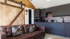 A modern kitchen with dark cabinets, a wooden wall, and a brown leather sofa with patterned cushions. - Romantic Countryside Lodge with Hot Tub (South Mundham)