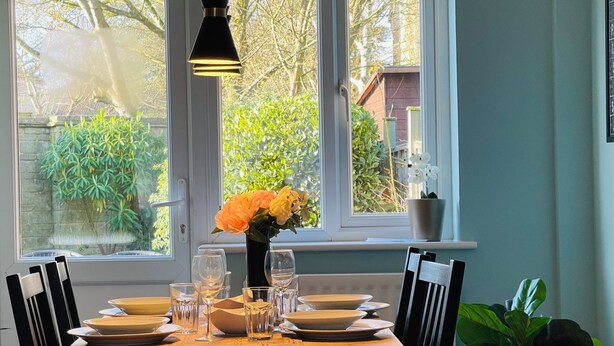 Ett matbord dukat med tallrikar, glas och en vas med blommor, placerat framför ett fönster med utsikt över grönska och ett hus.