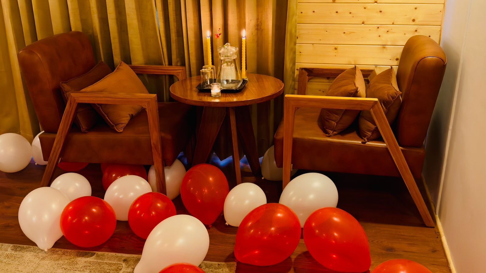 A room with two leather armchairs, a small round table with candles, and balloons on the floor.