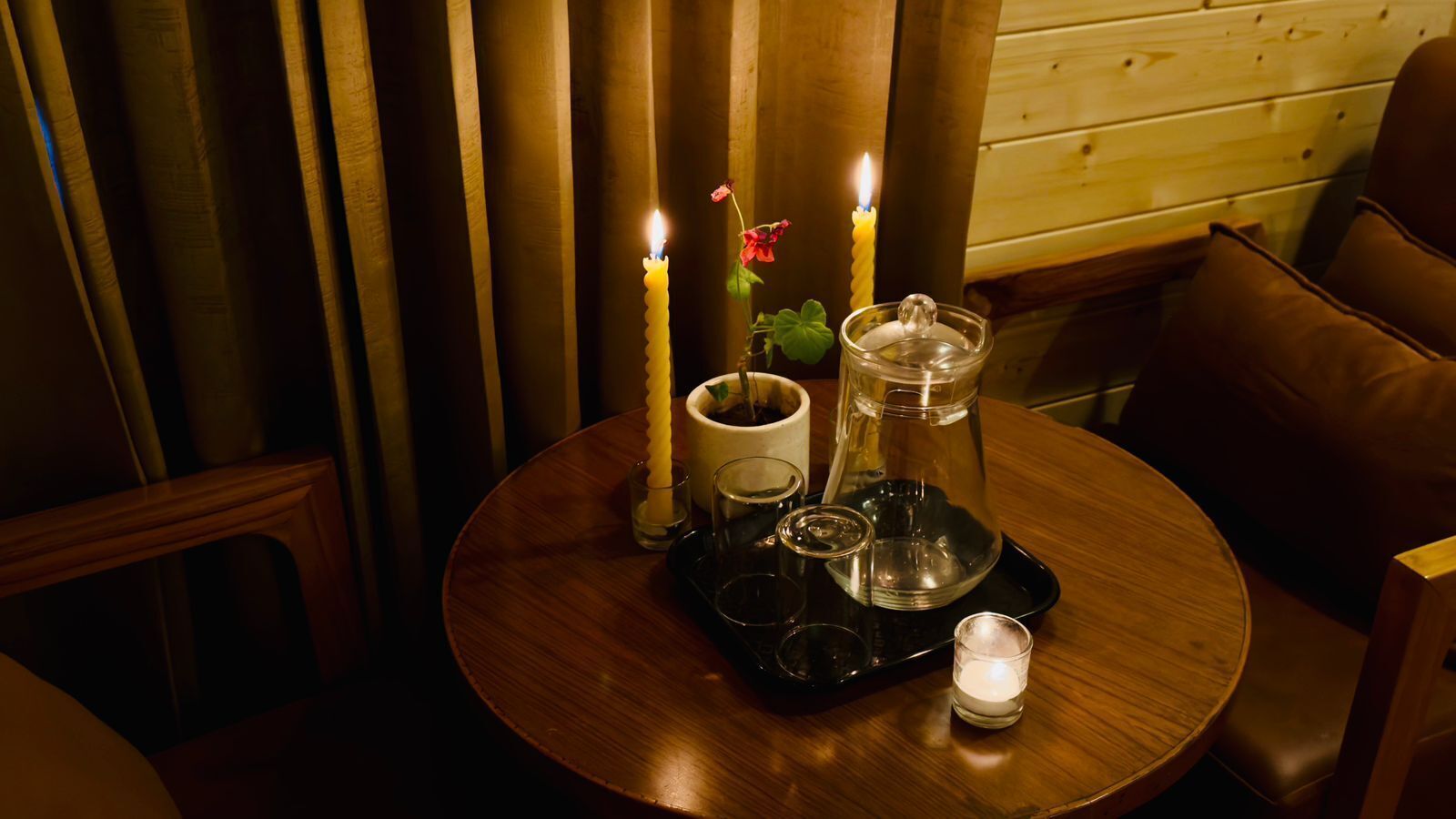 Een ronde houten tafel met kaarsen, een glazen kan en een dienblad met glazen en een kop.