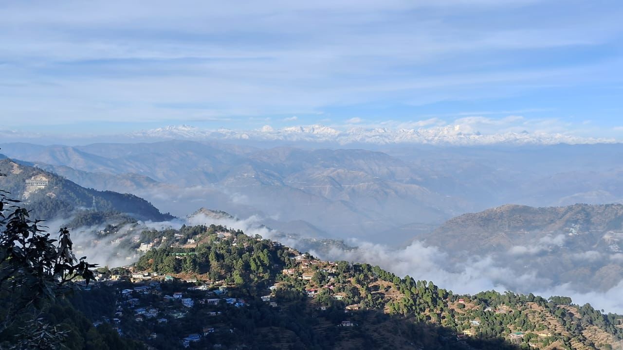 A mountainous landscape with a village nestled among the hills, surrounded by clouds and distant snow-capped peaks.
