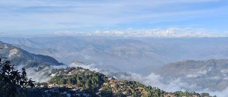 A mountainous landscape with a village nestled among the hills, surrounded by clouds and distant snow-capped peaks.