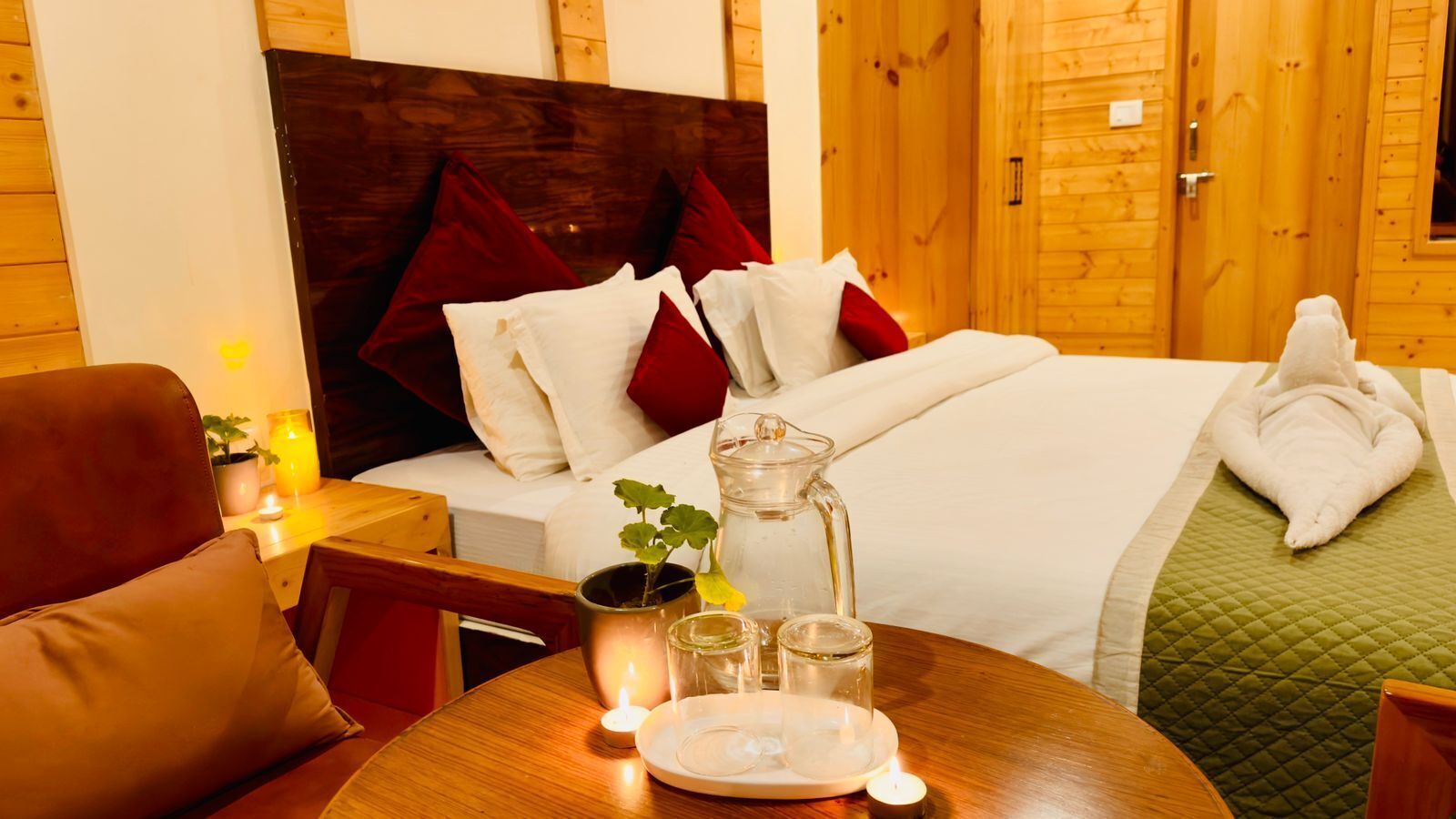 A cozy hotel room with a bed, a bedside table with candles, a vase with flowers, and a small table with glasses and a plate.
