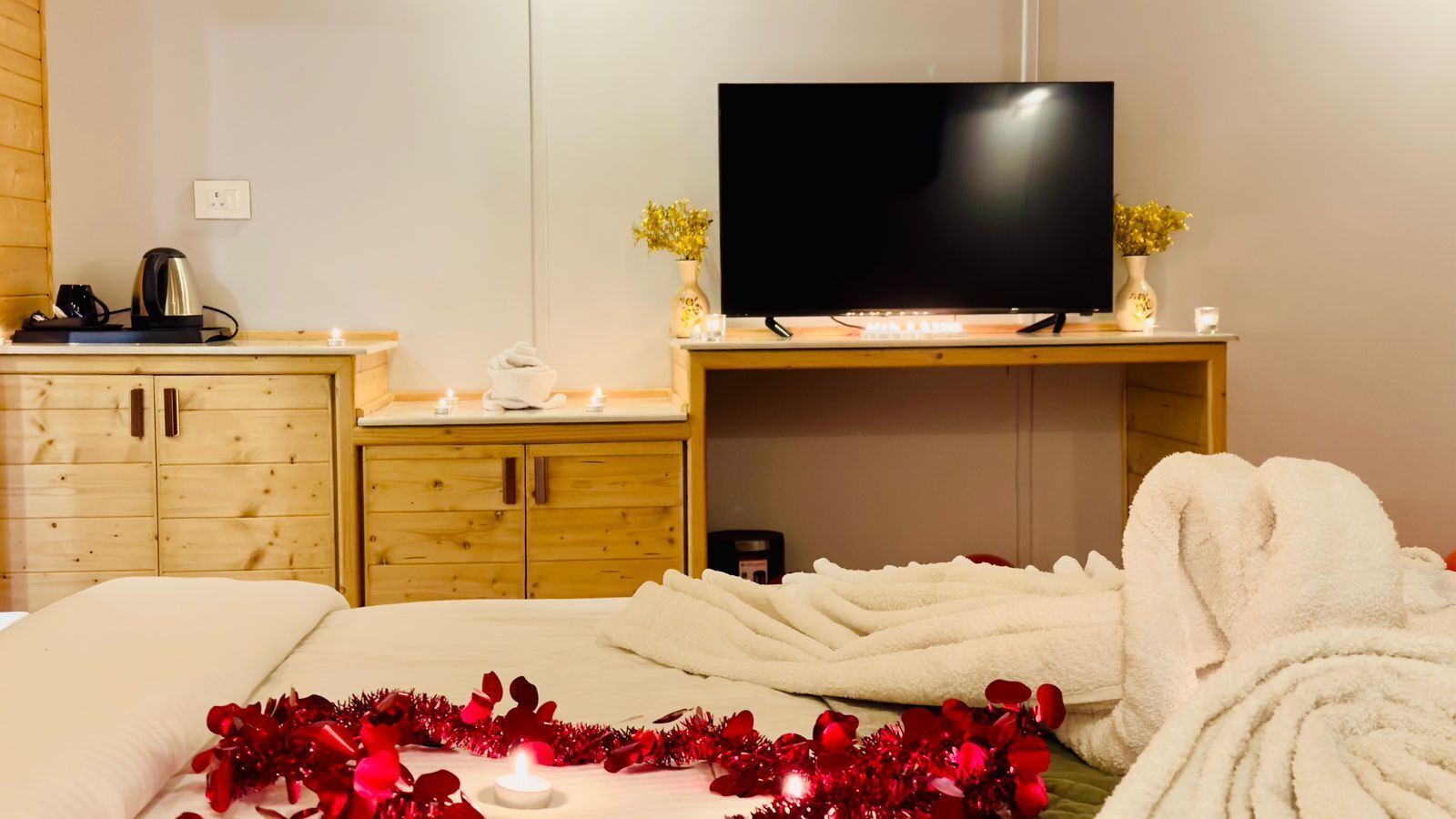 Une chambre d’hôtel avec un lit, un téléviseur posé sur un meuble en bois, une armoire en bois et une petite table sur laquelle se trouvent une bouilloire et un vase de fleurs.