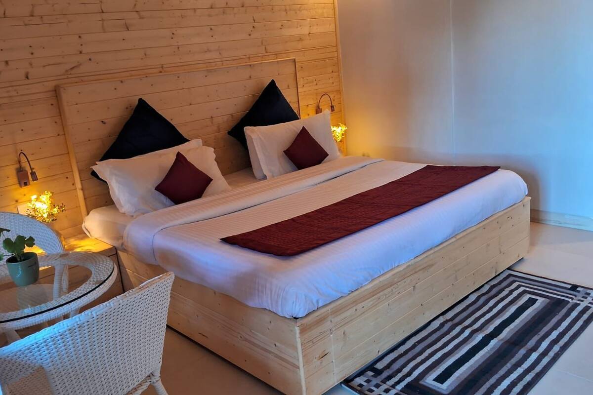 A bedroom with a wooden headboard, a bed with white bedding and red accents, a small round table with a plant, and a white wicker chair.