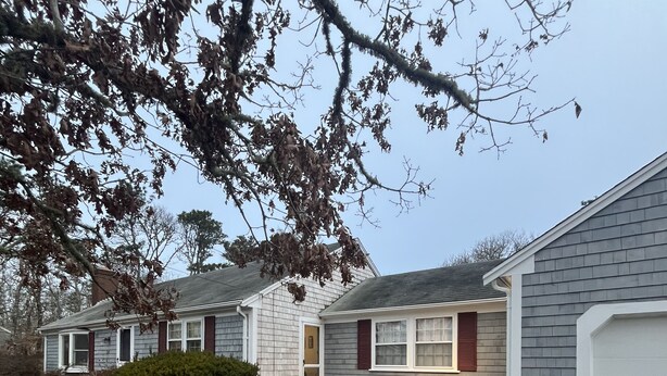 A residential property with a gravel driveway, a front yard with a small bush, and a house with a grey siding and red shutters.